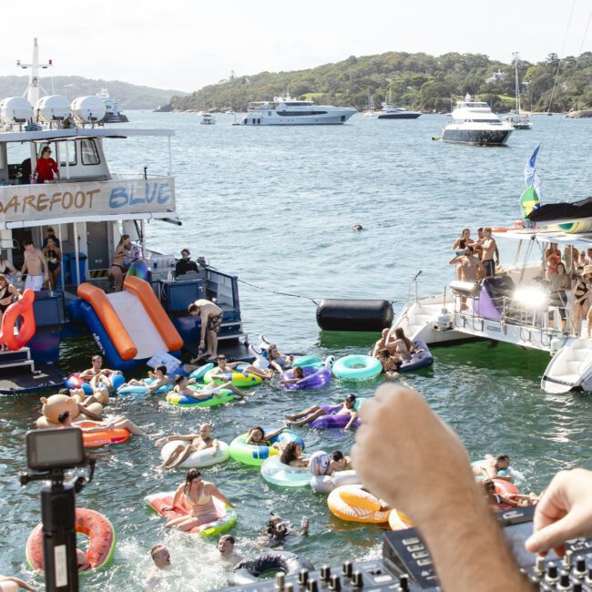 A DJ plays music while people enjoy a boat party on the water. Attendees are on colorful inflatables and boats, including one named "Barefoot Blue" with slides. The scene is lively, set against a backdrop of calm water and distant trees.