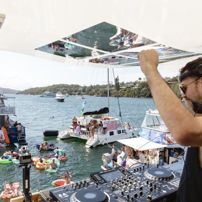 A DJ wearing sunglasses and headphones is playing music on a boat with DJ equipment set up. In the background, people are lounging on floaties and boats in the water, enjoying a sunny day. The scene is lively and festive.