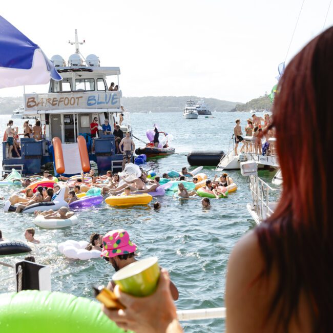 A lively scene with people swimming and lounging on colorful inflatables in the water, near a boat named "Barefoot Blue." Others are standing on the deck, while a woman in the foreground holds a drink. The background shows hills and more boats.