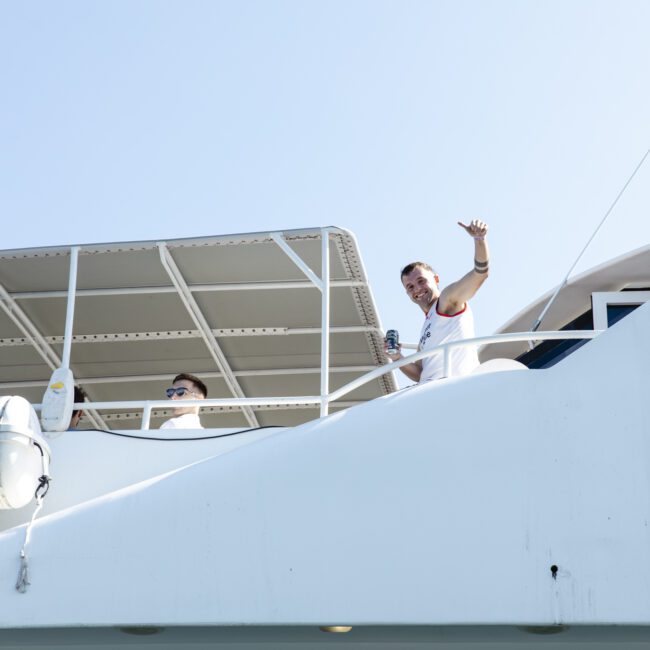 A person waves from the deck of a white yacht against a clear blue sky. Another person is partially visible next to them, wearing sunglasses. There are life buoys attached to the side of the yacht.
