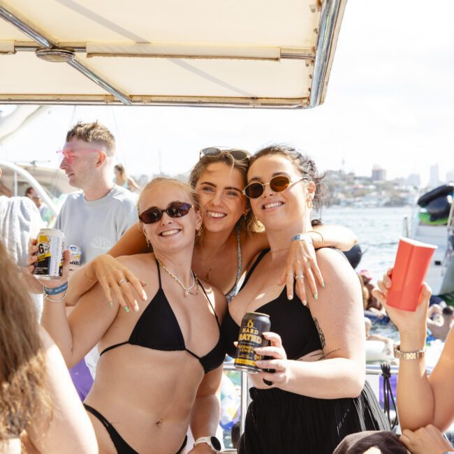 Three women in swimsuits pose happily on a boat, holding drinks. Two wear sunglasses, and they are smiling under a sunny sky. A man is visible in the background, and the scene is festive, with water and boats in the backdrop.