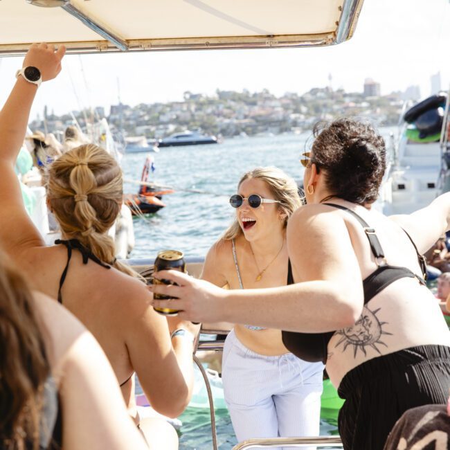A group of people enjoying a boat party under a canopy. One person with sunglasses in the center is laughing while another raises an arm, holding a drink. The background features a scenic view of water and distant land.