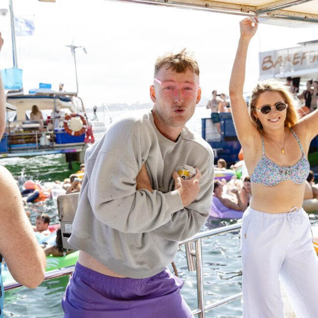 A person wearing a gray sweatshirt and purple shorts holds a can, posing playfully on a boat. Another person in a bikini top and white pants raises an arm joyfully. The background shows a lively party scene with people and boats on the water.