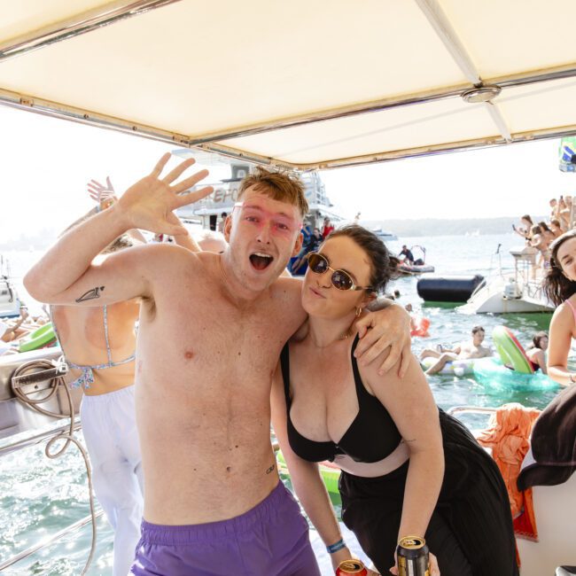 A group of people enjoying a boat party on a sunny day. A man and woman in the foreground smile and pose for the camera, with the man waving. Many others are in the background swimming and socializing. Floating devices and other boats are visible on the water.