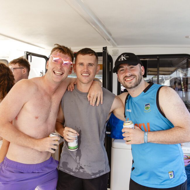 A group of friends on a boat, with three men in the center smiling at the camera. One man is shirtless wearing pink sunglasses, another in a gray tank top, and the third in a blue sleeveless shirt and cap. They are holding drinks.