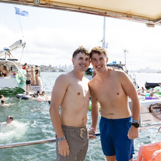 Two men smiling and posing on a boat with a festive atmosphere. People are swimming and enjoying inflatables in the water nearby. Other boats and a city skyline are visible in the background.