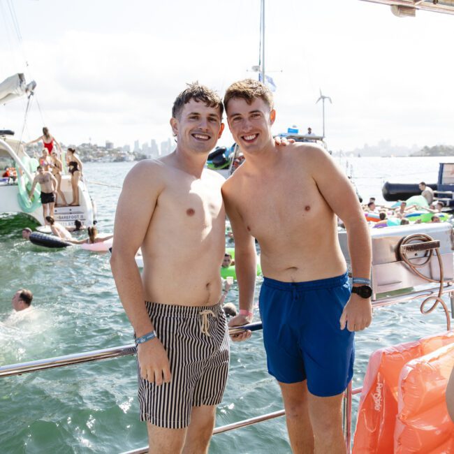 Two men in swim trunks stand on a boat, smiling for the camera. The background shows a busy harbor with other boats and people enjoying the sunny day on the water.
