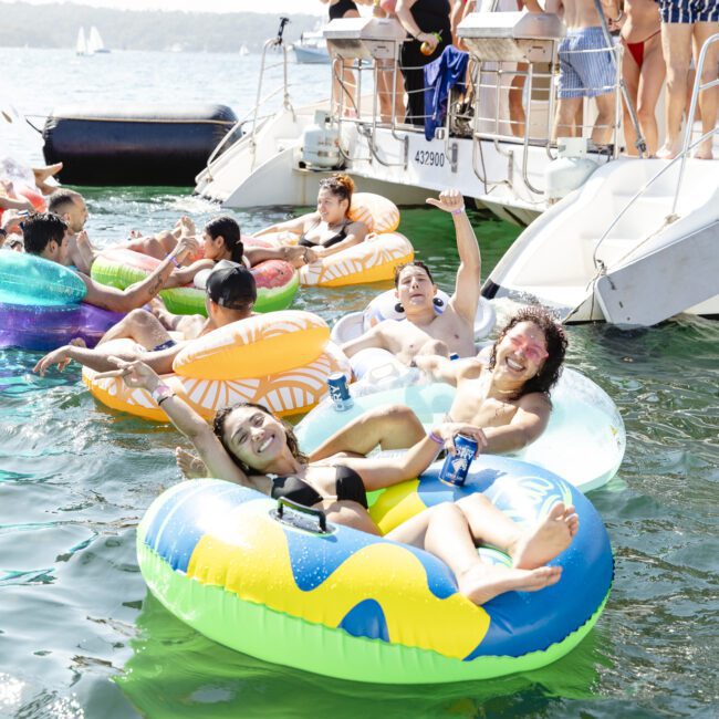 A group of people relax and have fun on colorful inflatable floats in the water near a yacht. Some hold drinks and smile, while others swim around. The sun is shining, and the atmosphere is lively and joyful.