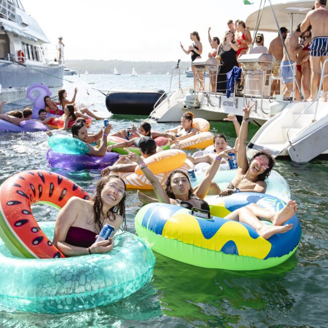 A group of people enjoying a sunny day on a lake, floating on colorful inflatable rings, including a watermelon and a snake pattern. Behind them, others stand and relax on a boat. The mood is festive and lively.