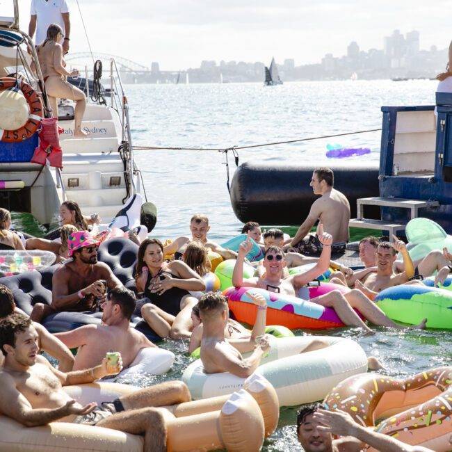 A lively group of people enjoying a party on inflatable floats in a harbor. Surrounding them are boats, and the city skyline is visible in the background. Some hold drinks, and the weather is sunny and clear.