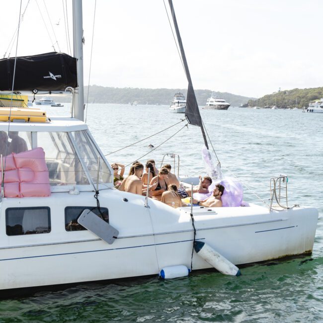 A group of people relaxes on a white sailboat with a black mast, anchored in calm waters. The sky is clear, and several boats are visible in the background. The shoreline with trees can be seen in the distance.
