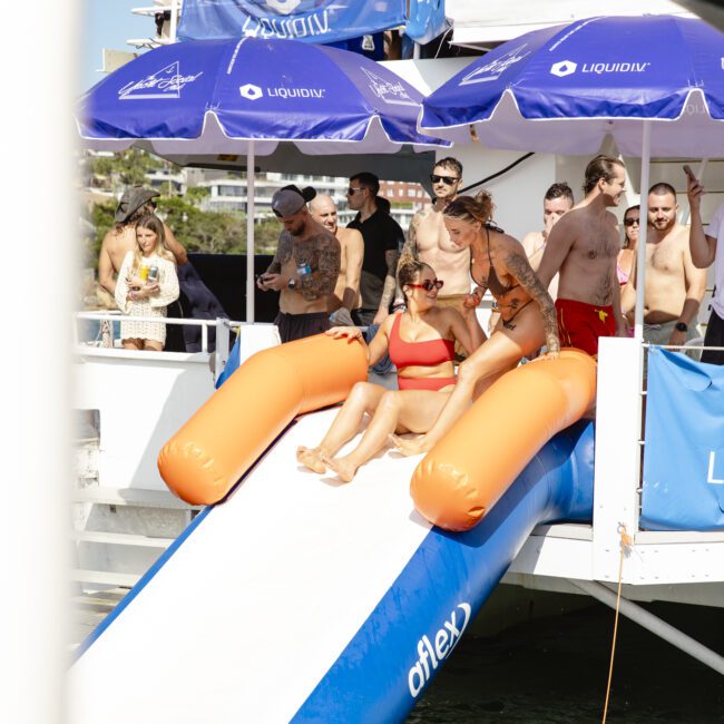 A group of people enjoying a sunny day on a boat. Some are sitting on an inflatable orange and white slide that leads into the water. Others are standing or sitting under blue umbrellas. The scene is lively and relaxed.