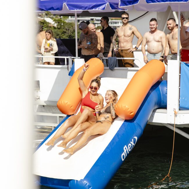 Two women in swimsuits slide down a large inflatable slide into the water, surrounded by people enjoying a sunny day. Others stand watching under purple umbrellas on a boat deck in the background.