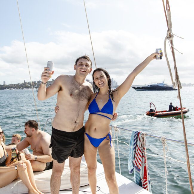 A man in swim shorts and a woman in a blue bikini stand on a boat, both holding drinks, smiling, and posing with arms raised. A group of people relax in the background near the water on a sunny day.