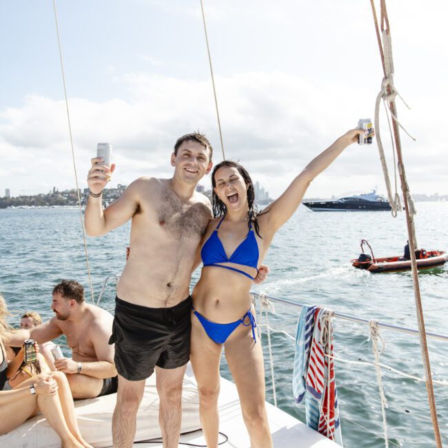 A shirtless man and a woman in a blue bikini pose and smile on a boat, each holding a drink. They are in front of the ocean with a city skyline visible in the background. Other people are seated on the boat, enjoying the sunny day.