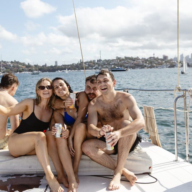 A group of friends sitting on a boat, smiling and holding drinks. They are wearing swimwear. The background features a sunny sky, water, and a distant shoreline with city buildings.