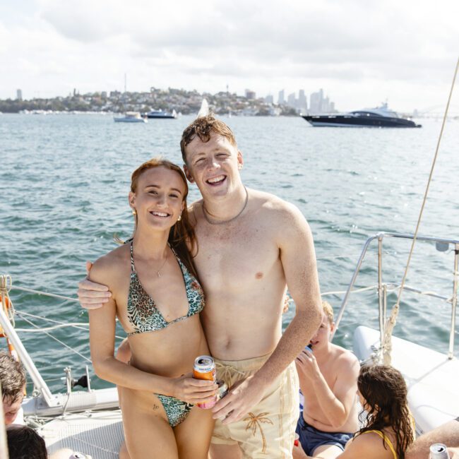 A group of people is relaxing on a boat. A man and woman in swimsuits stand smiling, with the man holding a drink. Other people are seated nearby. The background shows water and a distant shoreline with buildings under a partly cloudy sky.