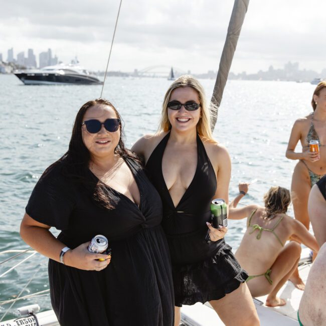 Two women in black dresses stand smiling on a boat with drinks in hand. They are surrounded by water with a city skyline in the background. Other people are on the boat, some in swimwear. It's a sunny day.