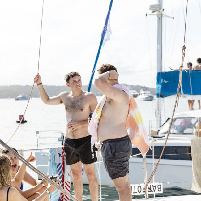 Two men in swim trunks stand on a boat, with one holding a towel. Several people are seated nearby, enjoying the sun. Other boats and a calm sea are visible in the background.