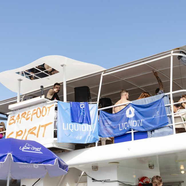A group of people enjoying a party on a yacht with banners reading "LIQUID IV" and one person DJing. An umbrella with the Liquid Death logo is visible. The sky is clear and blue.