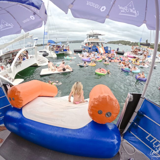 A lake party scene with people on inflatable floats and boats. A person sits on a large orange and blue floating structure under an umbrella. Large boats and a crowd enjoying the water are visible in the background. Overcast sky.