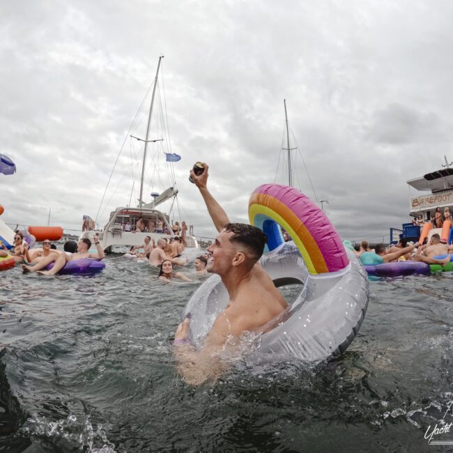 A man in a rainbow unicorn float raises a drink in a lively group gathering in a lake, surrounded by people in inflatable tubes, boats, and a cloudy sky.