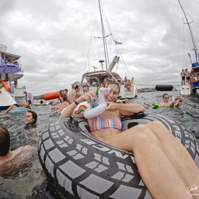 A woman in a colorful bikini floats on a large tire-shaped inflatable in the sea, surrounded by people swimming. Boats and a waterslide are in the background under a cloudy sky. The scene is lively and joyful.