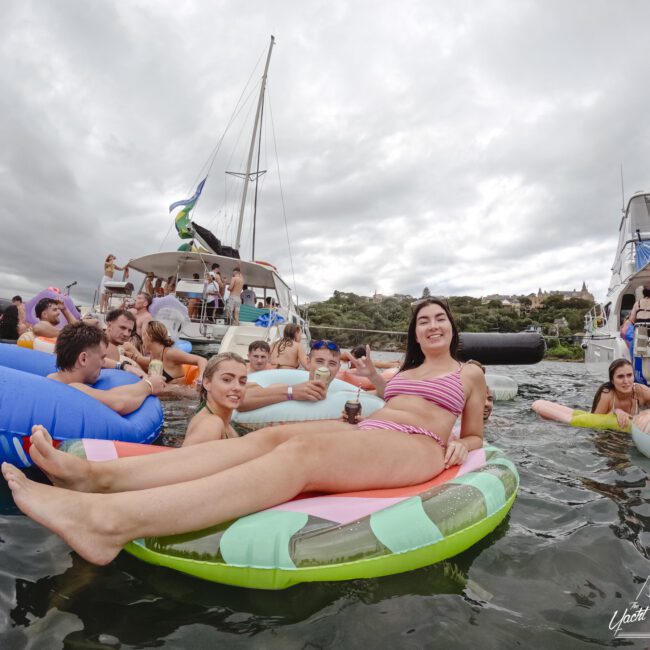 A group of people enjoying a party on a lake, lounging on colorful inflatables. A woman in the foreground is on a striped floatie, smiling. Several boats are nearby under cloudy skies, and others are socializing and relaxing.