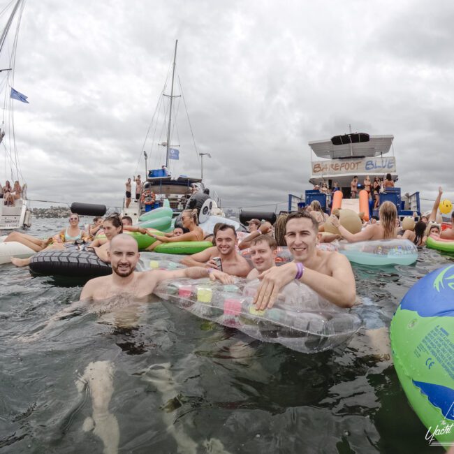 A group of people enjoying a party in the water with inflatable floats. There are boats in the background and a cloudy sky. The scene depicts a lively, fun atmosphere.