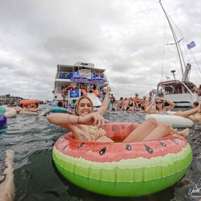 A person lounges on a watermelon-shaped float in a large body of water. Other people are in the water and on nearby boats in the background. The sky is overcast, and there's a festive atmosphere with flags on the boats.