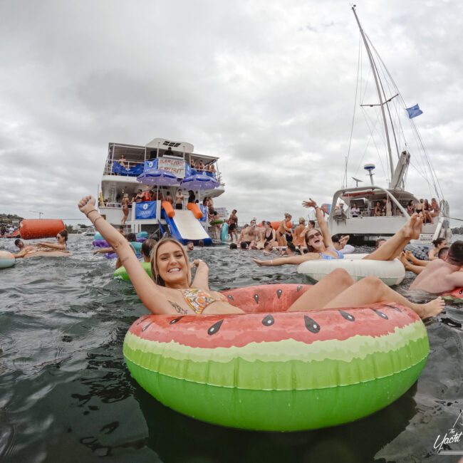 A person relaxes on a watermelon-themed float in a busy water scene. They are smiling at the camera. In the background, a yacht and slides are visible, along with many people enjoying the water. The sky is cloudy.