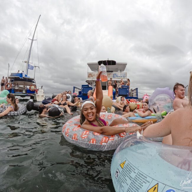 A group of people relax on inflatable tubes in the water near a boat. A smiling woman in a pink bikini raises a leg in the air while floating on a pizza-shaped tube. The sky is overcast, and the boat displays a "Bucket List Bash" sign.