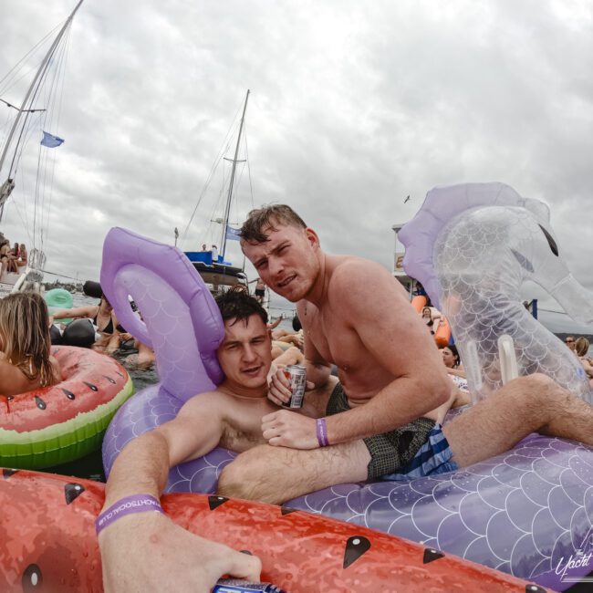 Two people are lounging on inflatable pool floats shaped like fruits in a crowded water setting. They are holding drinks and surrounded by others on similar floats. Boats are visible in the background under a cloudy sky.