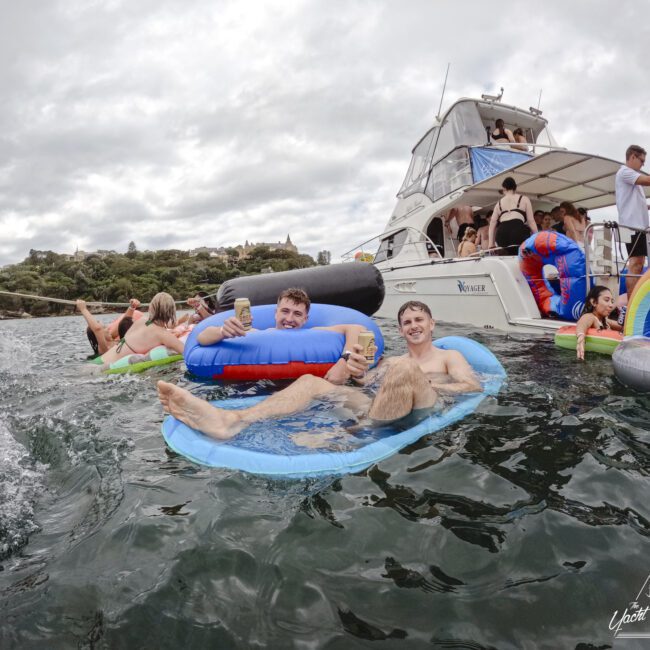 Two men float on inflatable loungers, holding drinks, near a boat with people on board. The water is wavy and overcast skies loom above. More inflatables are nearby, suggesting a relaxed, festive gathering.