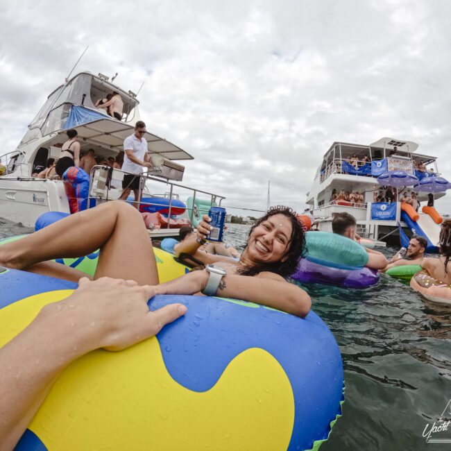 A woman smiles and lounges on a colorful inflatable in the water, surrounded by others on floats. Two boats with people are in the background. The sky is cloudy, creating a relaxed, lively atmosphere.