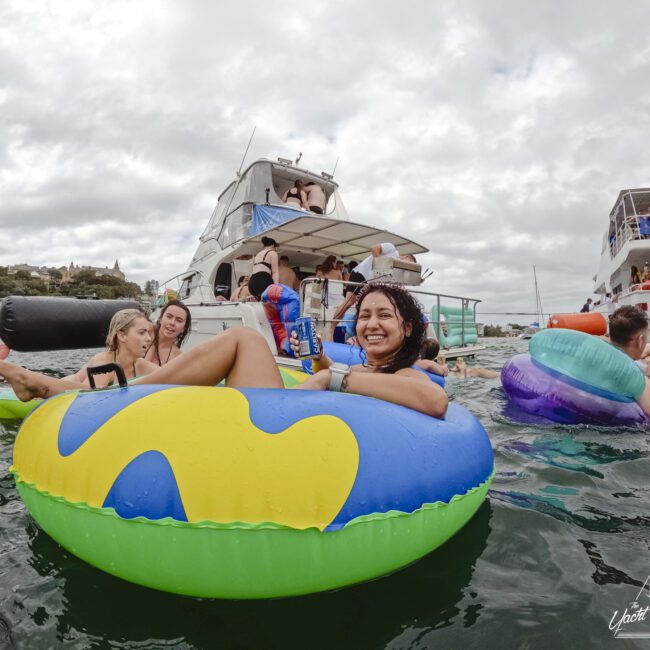 People are enjoying a day on the water, floating on colorful inflatables near a boat. The background features other boats and cloudy skies. Everyone appears relaxed and cheerful.