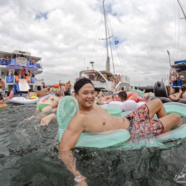 A man is lounging on a green inflatable float in the water, surrounded by other people on various inflatables. In the background, there are boats and more people, creating a lively, festive atmosphere under a cloudy sky.