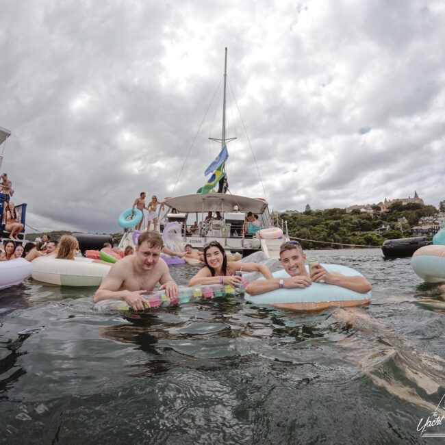 A group of people enjoying the water, smiling while holding inflatable rings near a boat. The boat is surrounded by other revelers, and there's a cloudy sky overhead. Trees and structures are visible in the background.