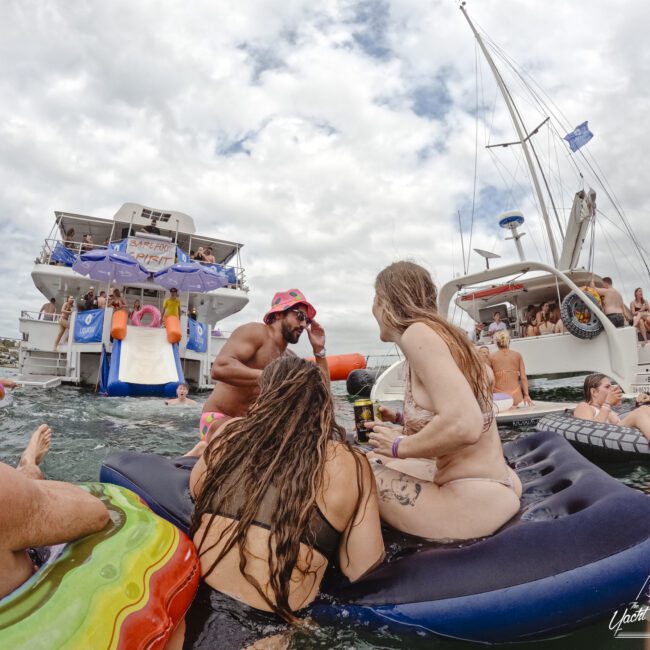 People in swimsuits enjoying a party on inflatable rafts and boats in the water. A large yacht with a slide is in the background, and a flag is flying on one of the boats. The sky is cloudy.