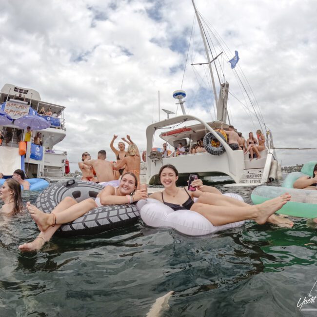 A group of people enjoying a boat party. Two women are floating on inflatable loungers in the water, surrounded by others on floats and in the water. Boats are in the background, and the sky is cloudy.
