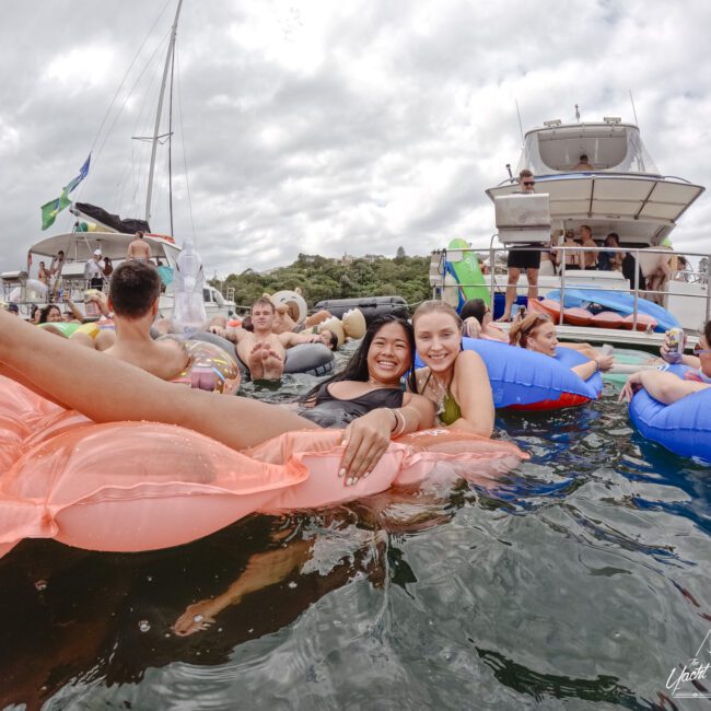 Two people smile while lounging on a pink float in water. Several others relax on blue floats nearby. Boats and more people are seen in the background under a cloudy sky, suggesting a festive atmosphere.