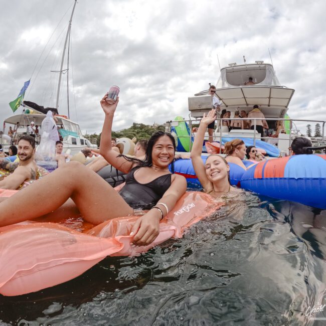 Two people smiling and holding drinks while relaxing on pool floats in the water. Boats and other people in the background indicate a lively and social atmosphere under a cloudy sky.