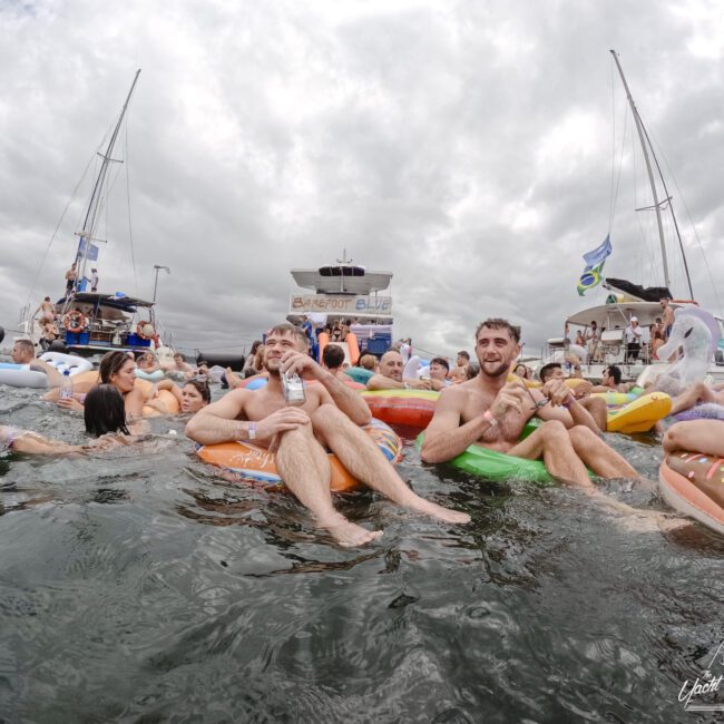 A group of people relax in the water on colorful inflatable rings. It's a cloudy day with two boats in the background. The scene is festive, with individuals enjoying themselves and one person holding a bottle.