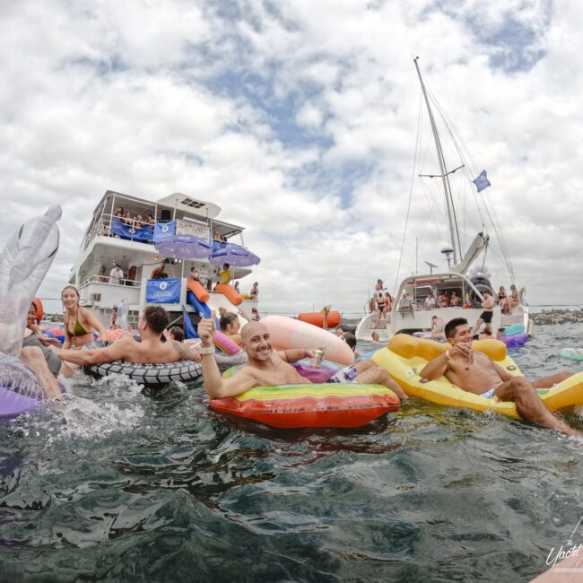 A group of people enjoy floating on colorful inflatables in the water near a large boat. They're smiling, holding drinks, and surrounded by more inflatables and another boat. The sky is cloudy.