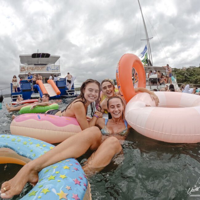 Three people smiling and posing with inflatable pool floats in the water. A boat with "Barefoot Live" signage and more people are in the background. The sky is cloudy and there are lush trees in the distance.