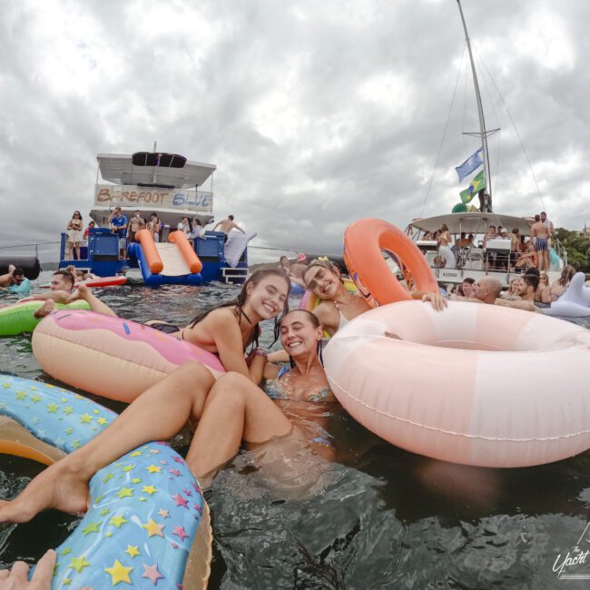 Three people smile while lounging on inflatable rings in the water near a boat named "Barefoot Blue." Other people and inflatables are visible in the background. The sky is cloudy, and there are trees on the shore.
