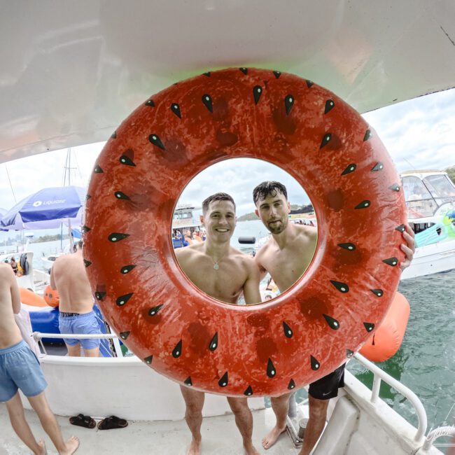 Two people smiling through a watermelon-themed inflatable ring on a boat. Others in swimwear are on the boat and in the water. Boats and trees are visible in the background under a cloudy sky.