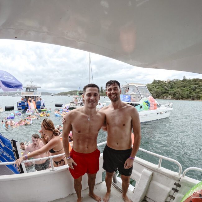 Two men in swim trunks smile on a boat. Behind them, people are enjoying a day in the water with inflatables. Several boats are anchored nearby. The sky is cloudy, and a green, hilly landscape is visible in the background.