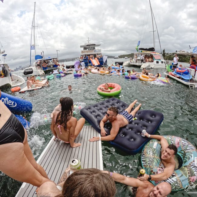People are enjoying a party on the water with colorful inflatable loungers. Several boats float nearby, and attendees hold drinks, swim, and relax under a cloudy sky. The atmosphere is lively and festive.