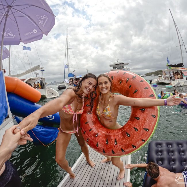 Two smiling women in swimsuits on a boat deck. One has a watermelon-shaped float. They're holding hands with someone off-camera. People and yachts are in the water around them, and a cloudy sky is overhead.